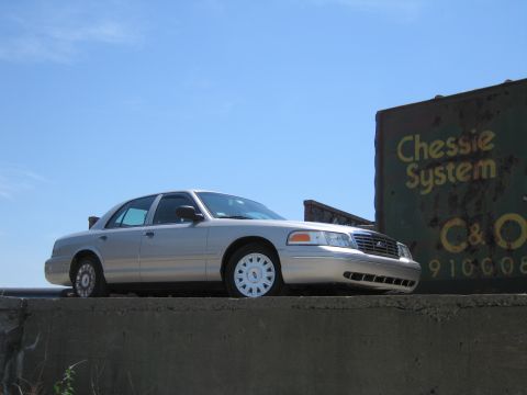 2001 Ford Crown Victoria - A silver 2001 Ford Crown Victoria is parked on a concrete ledge next to a weathered sign.