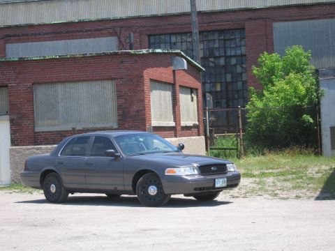 2001 Ford Crown Victoria - A gray Ford Crown Victoria is parked on a gravel lot in front of an industrial brick building.