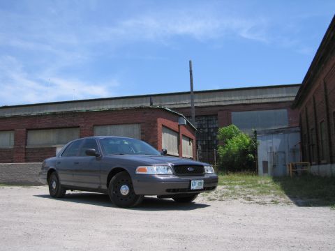 2001 Ford Crown Victoria - A 2001 Ford Crown Victoria sits parked on a gravel lot with industrial brick buildings in the background under a blue sky.