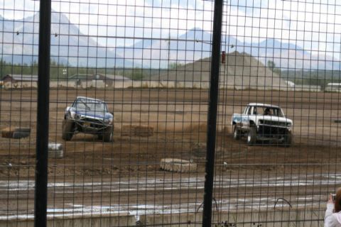 1975 Toyota Land Cruiser - Two modified trucks compete on a dirt track, kicking up dust as they race past the spectator area.