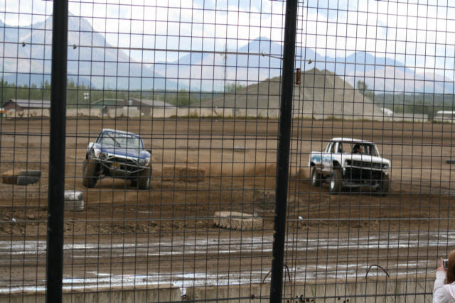 Two off-road trucks race on a dirt track with mountains in the background