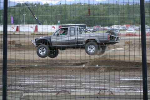 1975 Toyota Land Cruiser - A modified pickup truck is captured mid-air during an event, with a dirt track and spectators visible in the background.
