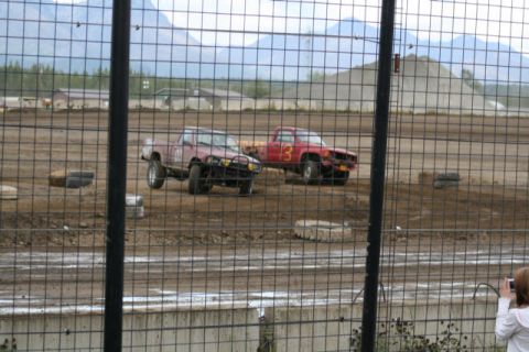 1975 Toyota Land Cruiser - Two modified pickup trucks compete on a dirt track, with mountains in the background.