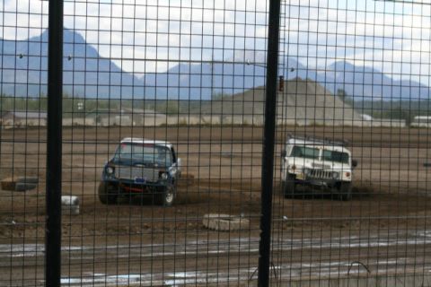 1975 Toyota Land Cruiser - Two vehicles compete on a dirt track, with a mountainous landscape visible behind a chain-link fence.