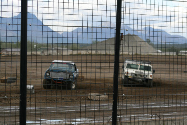 Demolition derby cars including white Hummer behind fence at dirt track