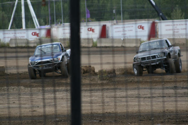 Two modified pickup trucks race on a dirt track, one blue and white, the other dark gray. Both have roll cages and off-road tires