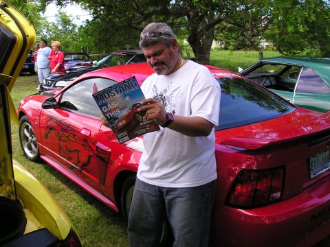 2009 Ford Mustang - Man reads Mustang Generations magazine next to a red Mustang with custom graphics at an outdoor car event.