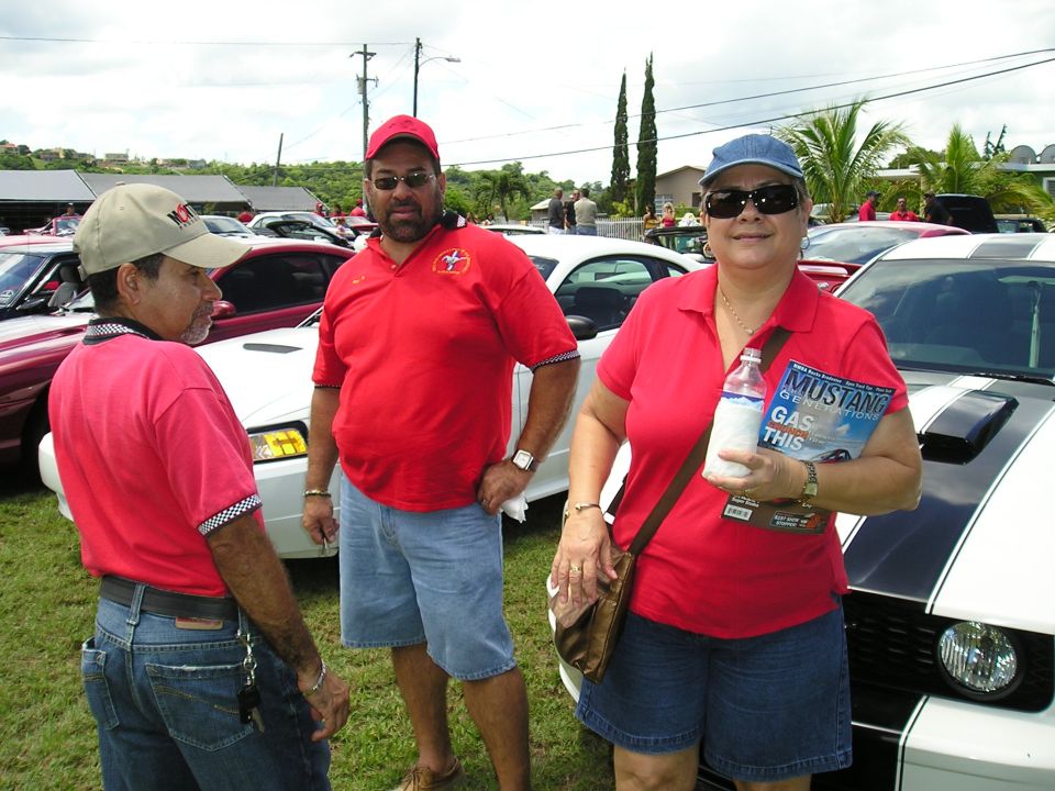 Three people in red shirts stand by a white Ford Mustang with black stripes. One person holds a Mustang magazine