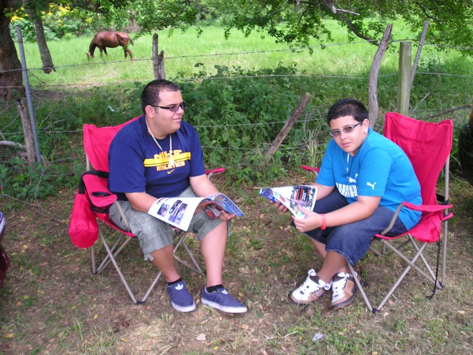 Two people sit in red folding chairs outdoors, reading magazines. A horse grazes in a field behind them