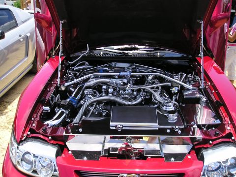 2009 Ford Mustang - Engine bay of a red car, extensive chrome detailing and a glimpse of another vehicle. The body style suggests a 2005 model year.