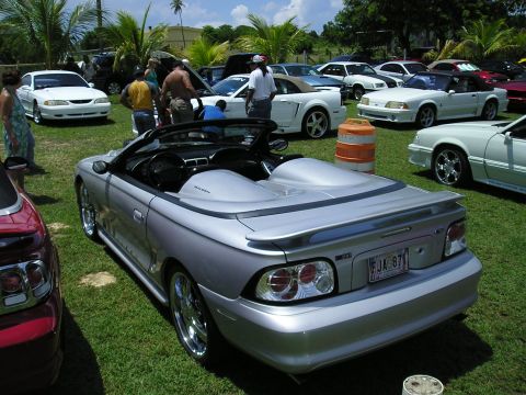 2009 Ford Mustang - A silver convertible Mustang is parked on grass with other vehicles and people around.