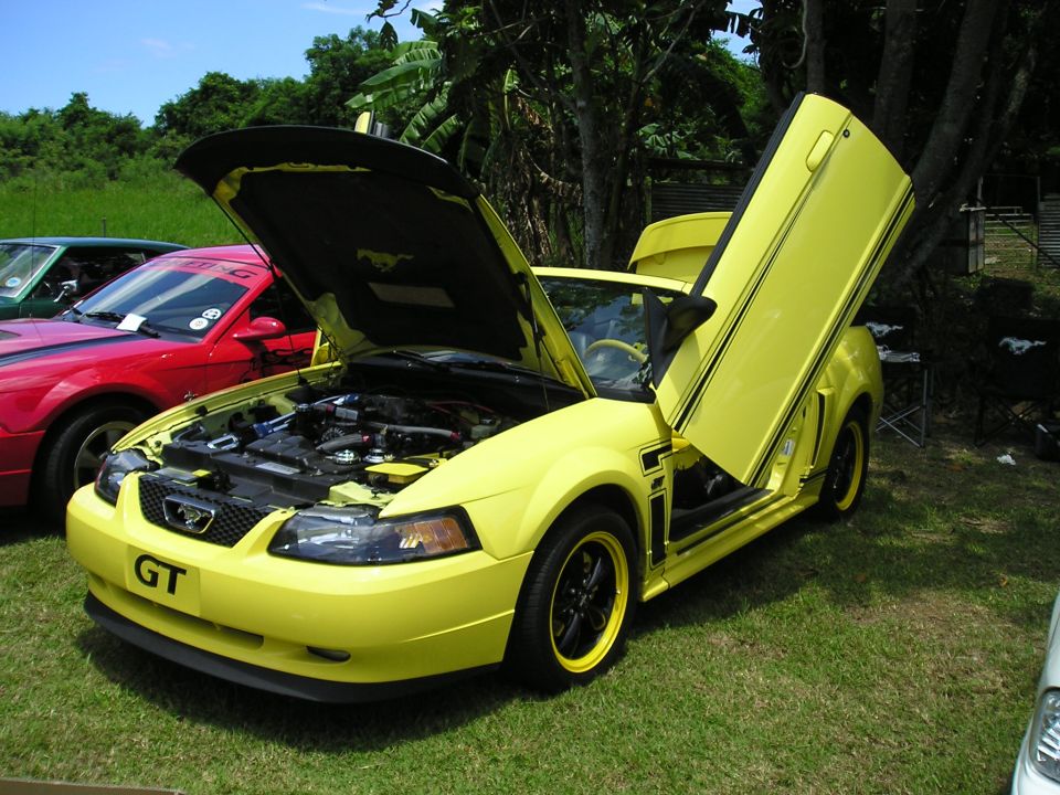 Yellow Ford Mustang with hood and lambo doors open, parked next to a red car
