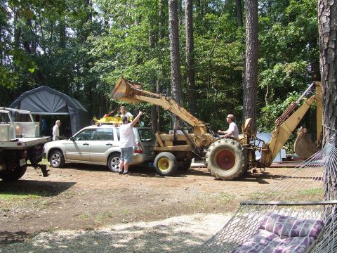 1967 Ford Bronco - A backhoe loader is positioned next to a silver SUV in a wooded area with several people present.
