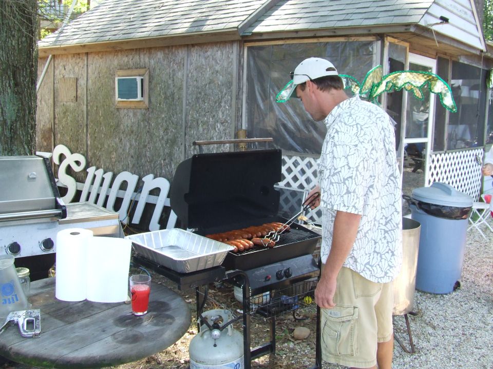 Man grilling hot dogs with dragonfly wings on his hat, rustic building in background.
