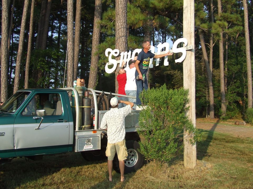 People installing a sign on a pole next to a green and silver pickup truck with propane tanks.