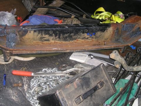 1989 Ford Bronco - Rusty steel truck bumper laid on its back in a cluttered cargo area, showing fresh weld beads at the end caps, bolt holes and blue-painted…