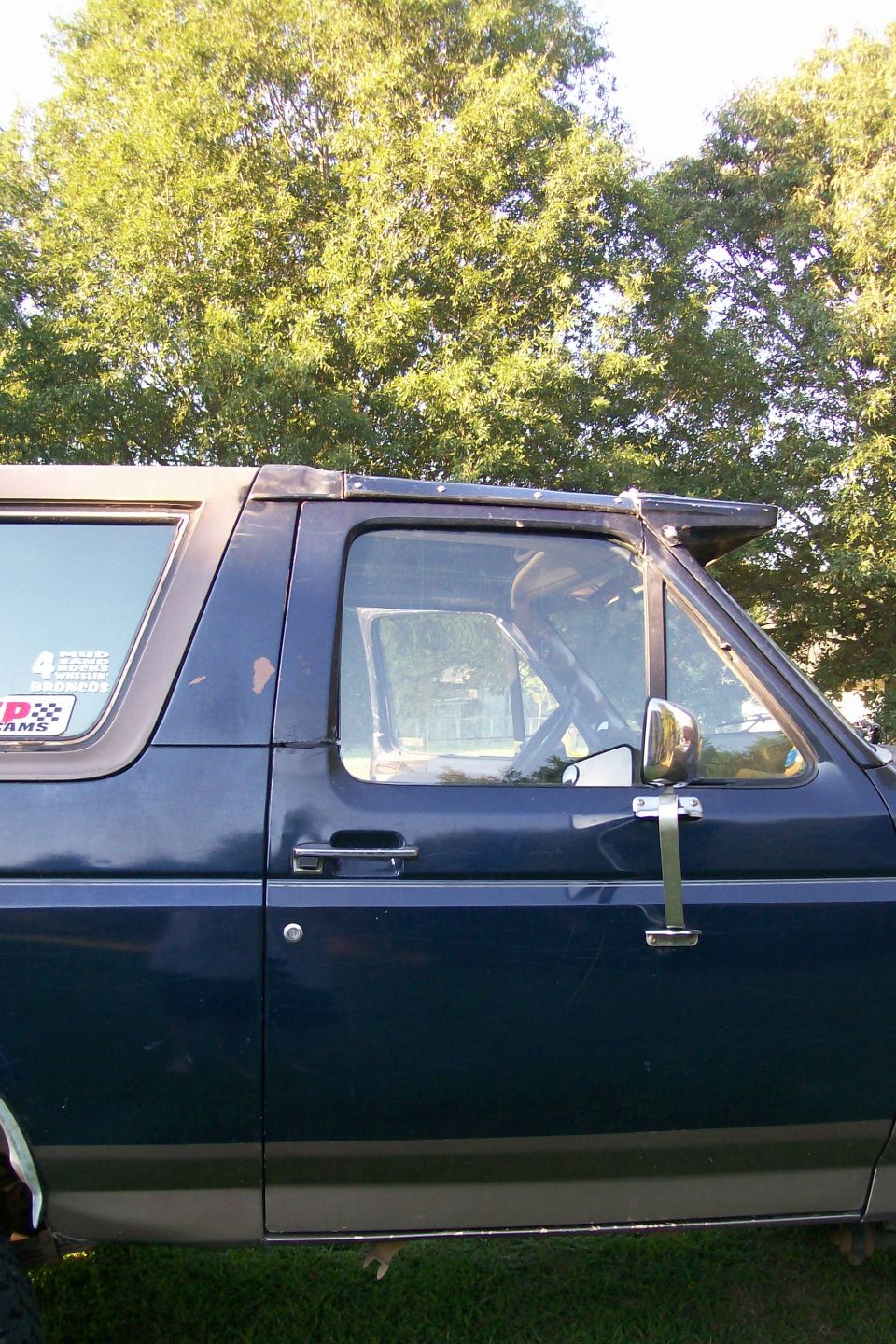 Dark blue two-tone Ford Bronco driver's door with chrome tow mirror and Broncos sticker, parked on grass
