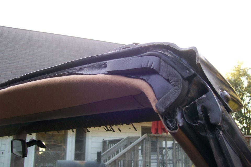 Close-up of windshield header corner showing weatherstrip seal and tan headliner trim on a black-framed vehicle