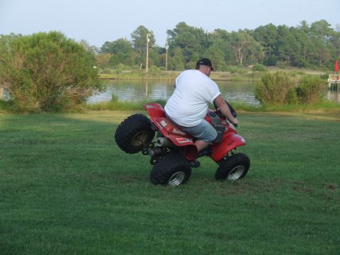 1967 Ford Bronco - A man rides a red ATV on its back wheels in a grassy field next to a lake.