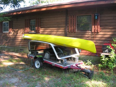 1991 Ford Bronco - A trailer is loaded with a yellow kayak and the disassembled frame of a vehicle, parked in front of a wooden house.