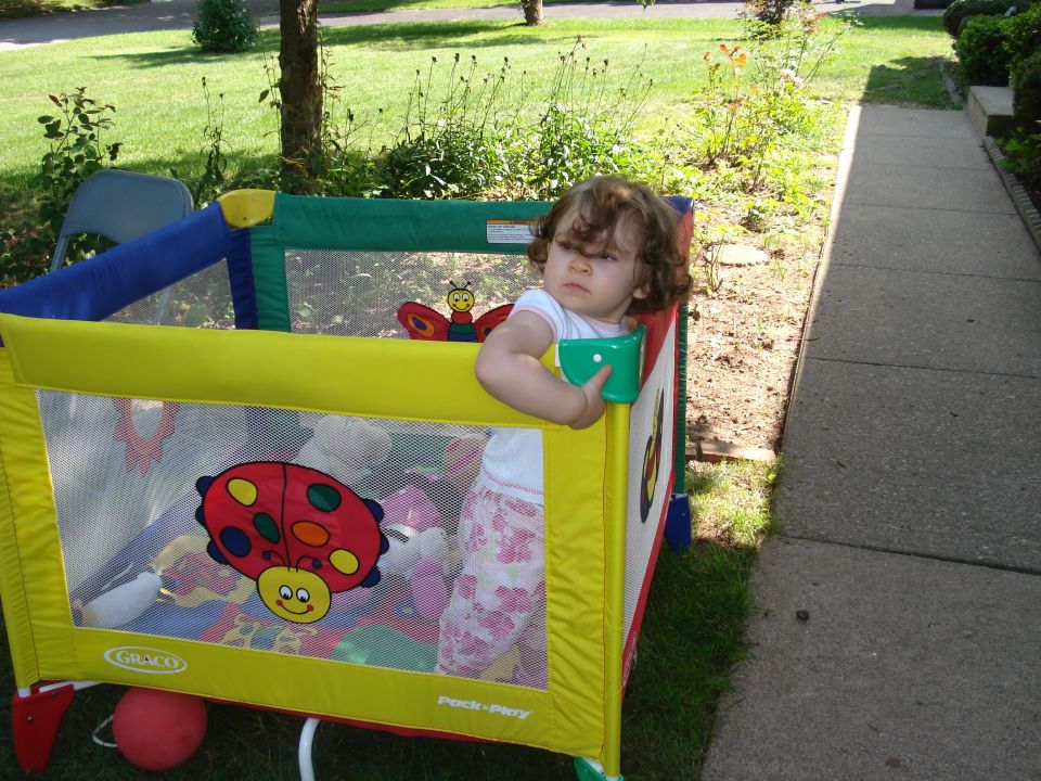 Baby in a yellow and green playpen with stuffed toys, outdoors