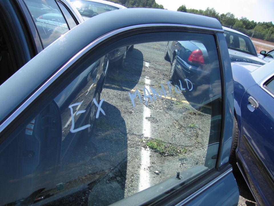 View through an open car door showing other vehicles parked in a lot. White writing on the window reads 'EX MANIFOLD'
