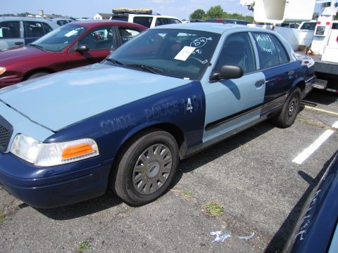 2001 Ford Crown Victoria - A two-tone blue police car is parked in a lot with other vehicles, displaying partial text on its fender.