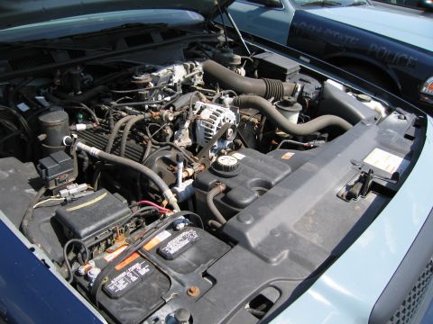 2001 Ford Crown Victoria - Engine bay of a 2001 Ford Crown Victoria, showing the battery and other components. A police vehicle is in the background.