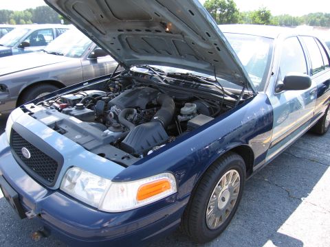 2001 Ford Crown Victoria - The engine compartment of a blue Ford Crown Victoria is visible with the hood raised, parked next to another vehicle.