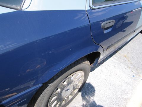 2001 Ford Crown Victoria - The rear side panel of a blue and light blue 2001 Ford Crown Victoria shows significant scratches and a dent.