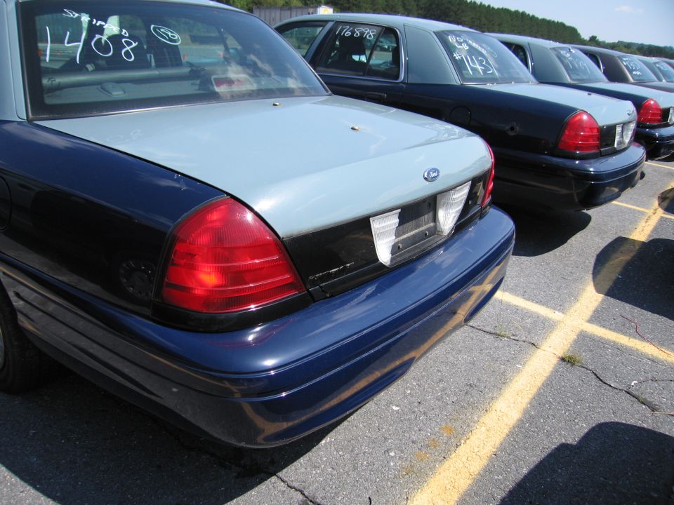 Rear of a blue and gray 2001 Ford Crown Victoria, parked in a lot with other similar cars. Numbers '1408' are written on the rear window
