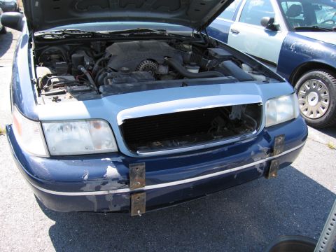2001 Ford Crown Victoria - The front of a blue 2001 Ford Crown Victoria is shown with its hood open, revealing the engine and a missing grille.