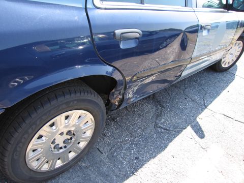 2001 Ford Crown Victoria - The rear passenger side of a blue sedan shows extensive damage, including rust and dents.