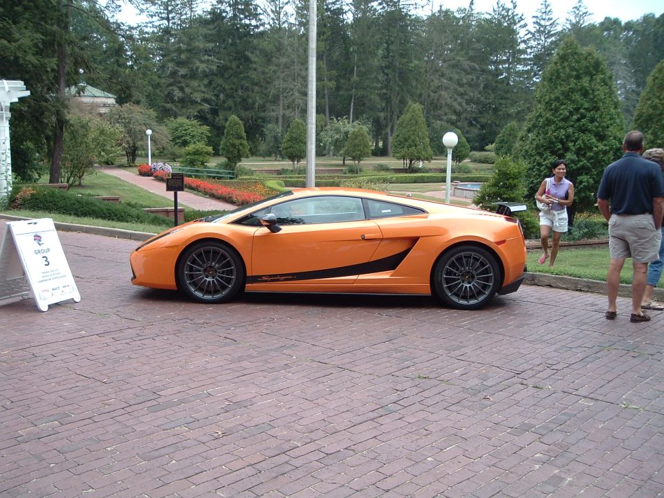 orange sports car parked on brick driveway, people standing nearby