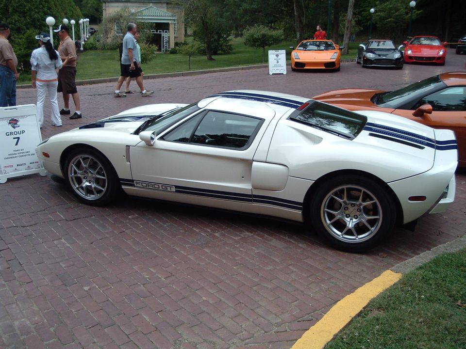 White Ford GT with blue stripes parked on brick pavement, other sports cars in background
