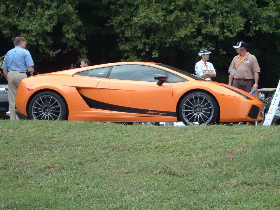 Orange sports car with black stripe on side. Text on side reads 'Superleggera'. People standing around it