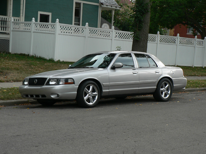 Silver full-size sedan with five-spoke chrome wheels parked curbside by a white fence