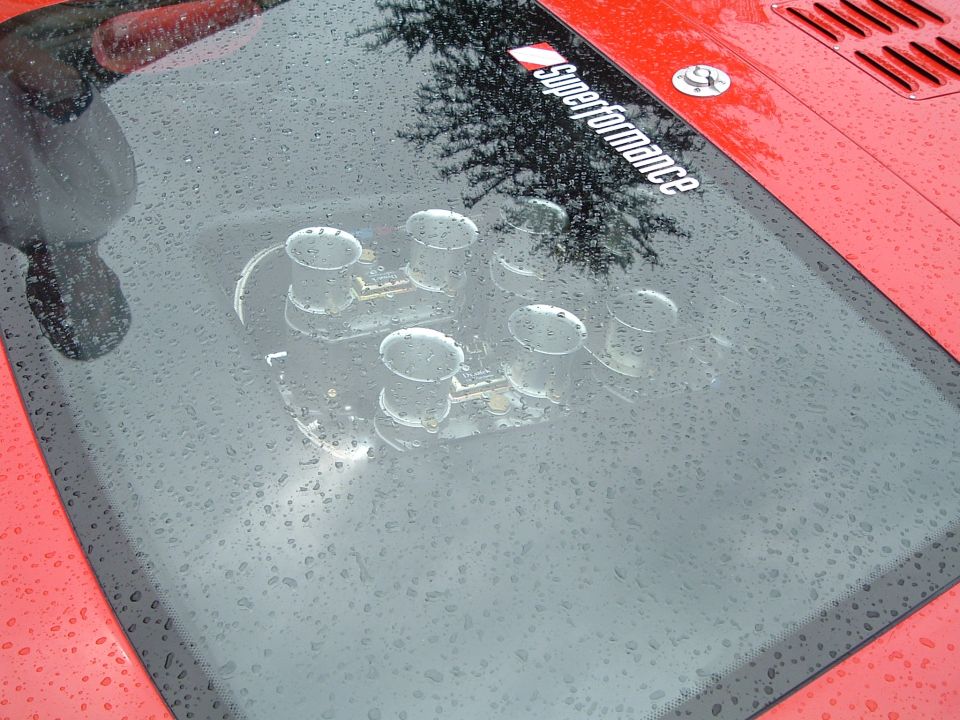 Red car hood with a black panel showing a six-cylinder intake manifold with water droplets