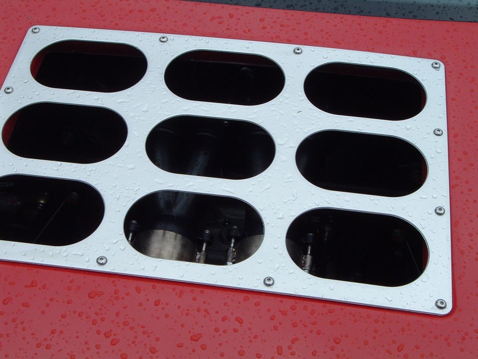 Silver engine intake manifold cover with nine oval openings, bolted to a red surface with water droplets