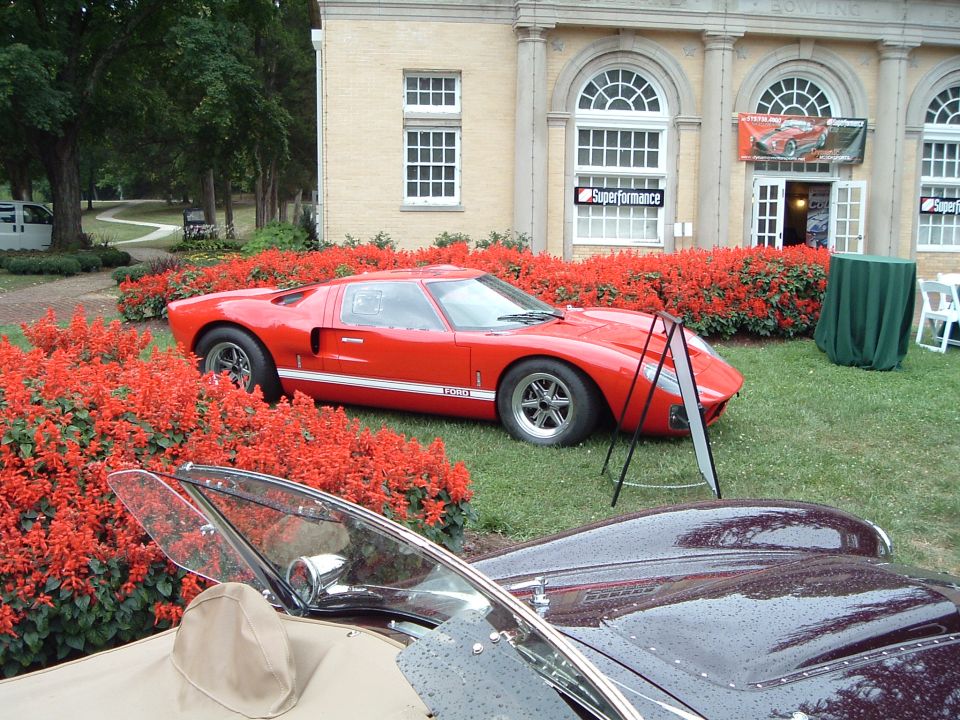 Red sports car with white stripe, parked near red flowers and a building. Another car's hood is in the foreground