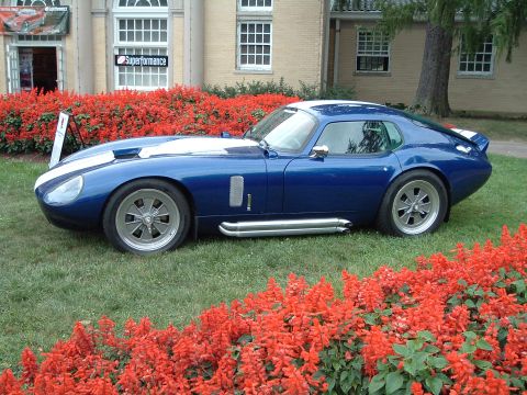 1999 Mercury Grand Marquis - A blue Shelby Cobra Daytona Coupe replica is displayed outdoors with red flowers in the foreground and a building in the background.