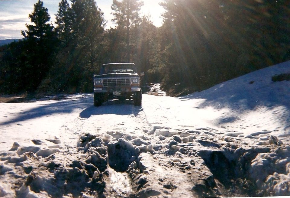 Lifted Ford Bronco facing camera on a snow-covered forest trail with sun flaring through pine trees