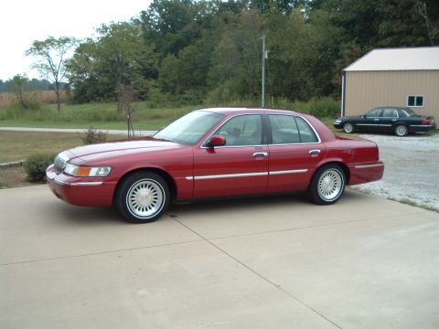1999 Mercury Grand Marquis - A red 1999 Mercury Grand Marquis sedan is parked on a driveway, with another dark sedan visible in the background.