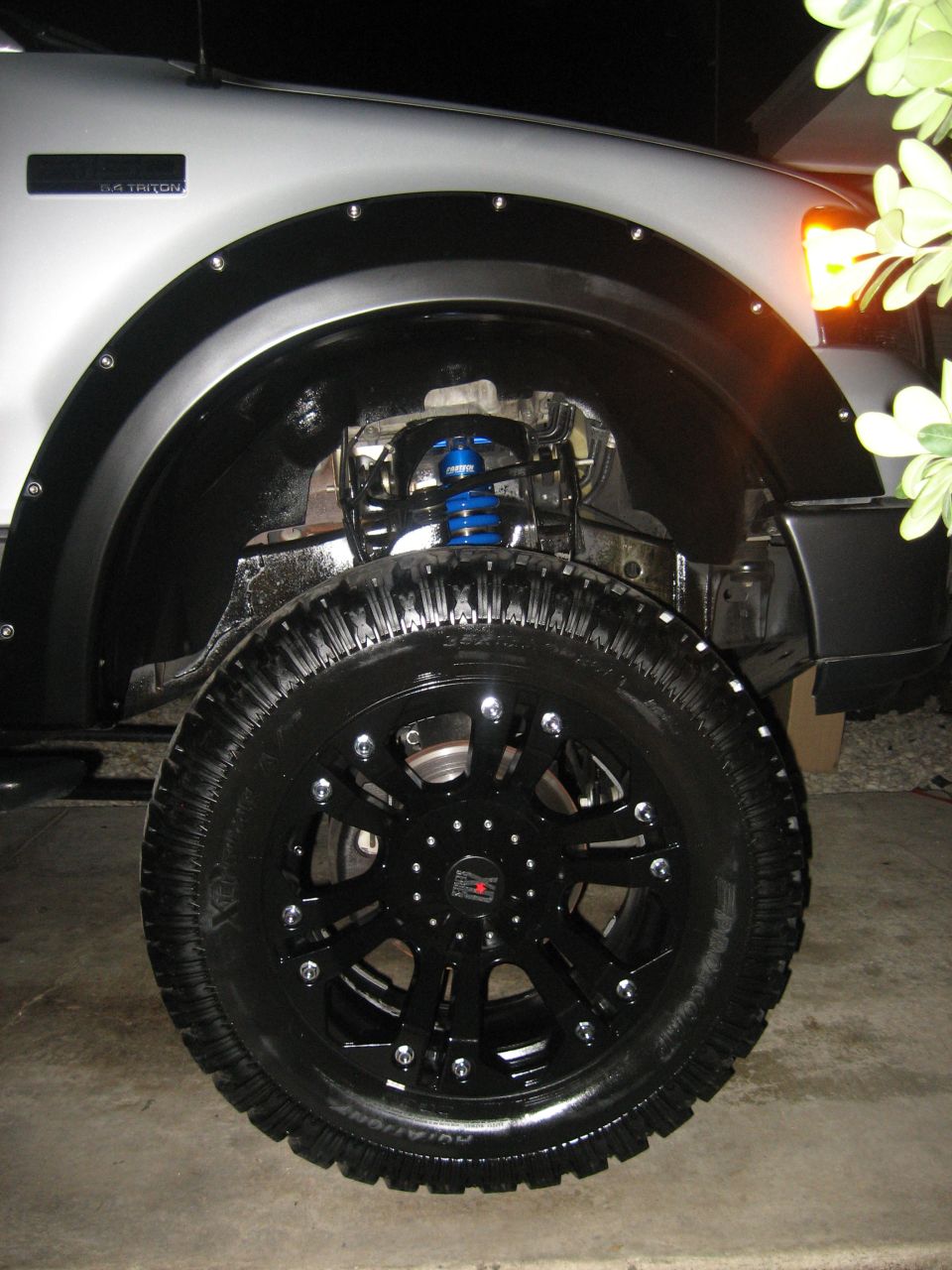 Silver truck fender with black fender flare, custom black wheel, and blue suspension spring