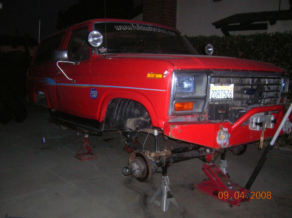 Red 1985 Ford Bronco on jack stands with front end disassembled, showing suspension and bumper details. License plate 2DRT526