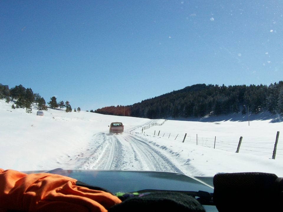 Driver's view following a tan truck on a snowy road through fenced winter fields with pine trees