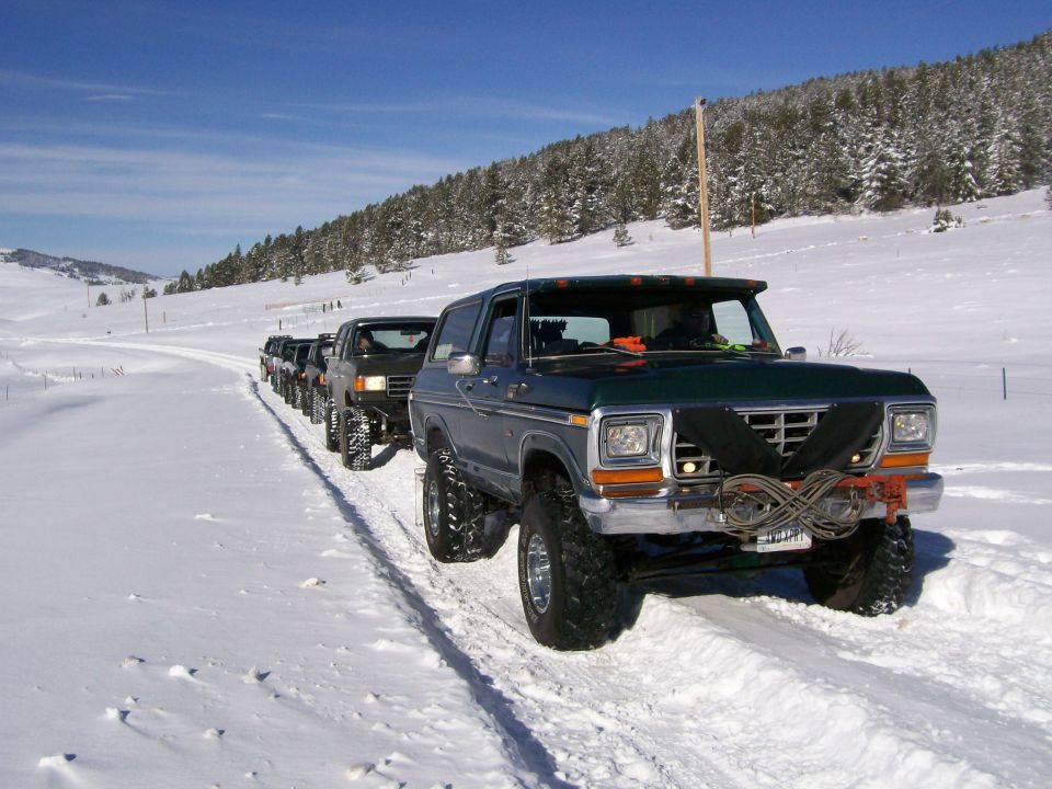 Lifted green Ford Bronco with big tires leading a convoy of trucks on a snowy trail
