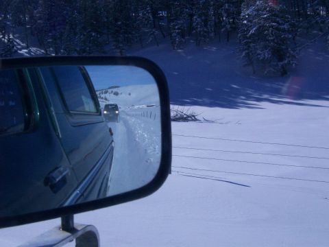 1979 Ford Bronco - A green SUV is seen from its side mirror driving on a snow-covered road with another vehicle ahead.