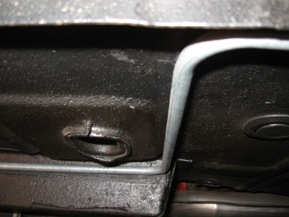 Underside of a vehicle showing a metal gas line running along the black undercoating near a cracked opening