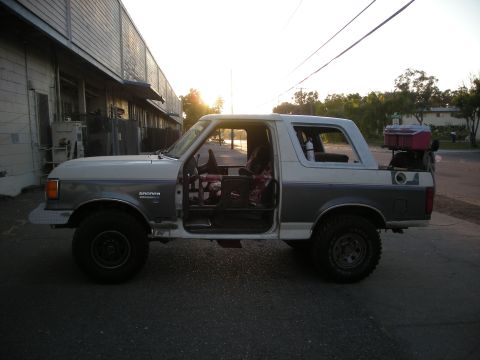 1991 Ford Bronco - This two-tone Ford Bronco is seen from the side with its doors off, parked in front of a commercial building during golden hour.